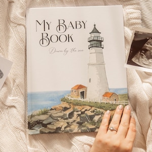 Puede incluir: Un libro blanco para bebés con el título "My Baby Book" y el texto "Down by the sea". La portada presenta una ilustración de acuarela de un faro en una costa rocosa.