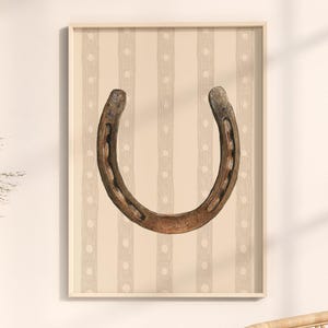 May include: A framed print featuring a horseshoe illustration. The horseshoe is brown and appears aged, set against a beige background with vertical stripes and dotted accents. The artwork is displayed in a light-colored wooden frame.
