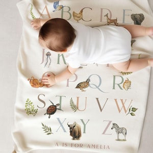 Puede incluir: Una manta de bebé blanca con un alfabeto colorido impreso en ella. Cada letra está emparejada con una ilustración de animal. La manta tiene el texto "A is for Amelia" en la parte inferior.