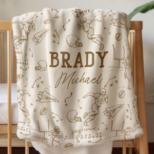 Könnte beinhalten: Cremefarbene Babydecke mit Football-Motiv. Die Decke zeigt ein sich wiederholendes Muster aus Footballspielern, Helmen und Spielschemata in Braun. Der Name "BRADY Michael" ist prominent in der Mitte zu sehen.