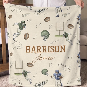 Puede incluir: Una manta color crema con un tema de fútbol, con ilustraciones de jugadores, cascos y balones de fútbol. La manta tiene el nombre "HARRISON James" impreso en marrón. El texto adicional incluye "FIRST DOWN" y "MVP".