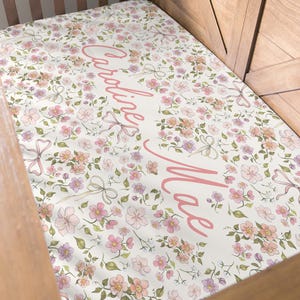 May include: A floral baby blanket with the name "Caroline Mae" in pink script. The blanket features a pattern of pink, peach, and purple flowers with green leaves and pink bows on a white background. The blanket is in a wooden crib.