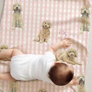 May include: A baby in a white onesie lies on a pink and white gingham patterned blanket. The blanket is decorated with illustrations of light brown dogs and pink bows. The baby's head and arms are visible.