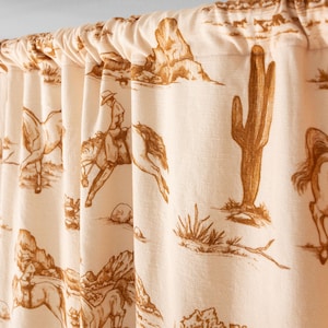 May include: A gathered curtain featuring a repeating toile-style print in brown ink on a cream-colored fabric. The pattern depicts desert landscapes with cacti, rocky formations, and figures of people riding horses in a western-themed scene.