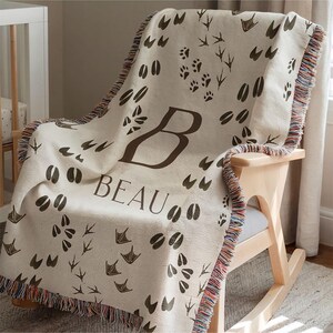 May include: A white woven baby blanket with a brown print of animal footprints and the name "Beau" in brown text. The blanket is fringed and has a woven texture.