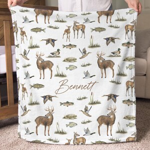 May include: White blanket with a repeating pattern of deer, fish, ducks, and cattails. The blanket features the name "Bennett" in a brown script font. The blanket is held up by a person.