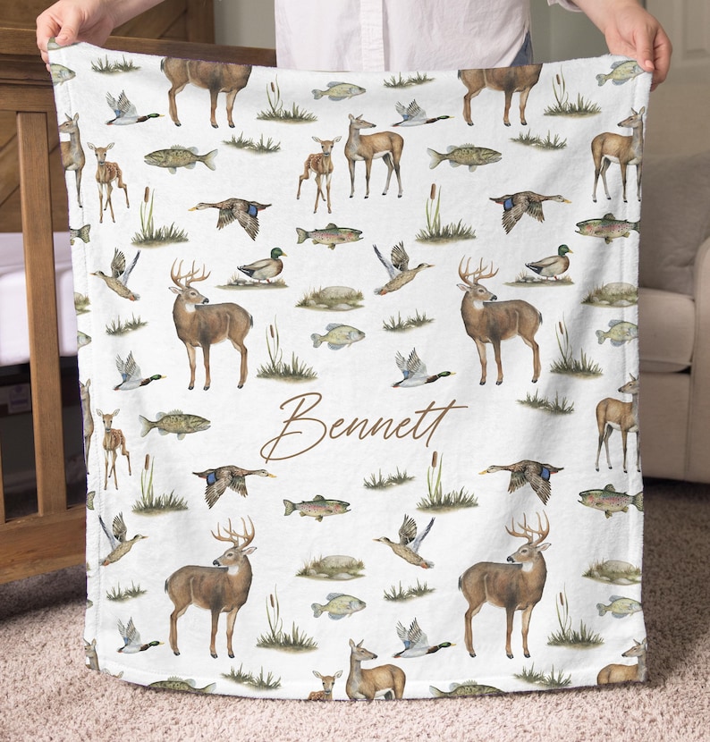 May include: White blanket with a repeating pattern of deer, fish, ducks, and cattails. The blanket features the name "Bennett" in a brown script font. The blanket is held up by a person.