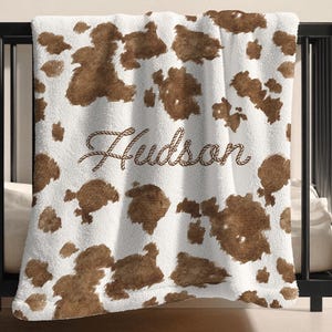 May include: White and brown cow print blanket with the name "Hudson" in a rope-style font. The blanket is draped over a black crib. The blanket has a soft, plush texture and a repeating pattern of brown cow spots.