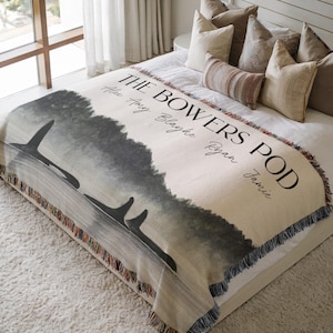 May include: A woven throw blanket featuring a watercolor-style illustration of orcas swimming in dark water. The text reads THE BOWERS POD followed by the names Alex, Amy, Blayke, Ryan, and Jamie. The blanket has a fringed edge and a cream background.