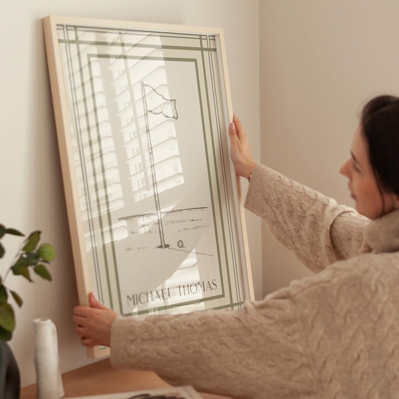 May include: A framed art print featuring a minimalist illustration of a flag on a pole, with the text "MICHAEL THOMAS" at the bottom. The artwork has a light beige frame and a green border. The image is being held up.