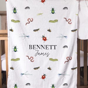 May include: White baby blanket with a repeating pattern of watercolor insects, including ladybugs, dragonflies, and bees. The name "BENNETT James" is printed in black script. The blanket is held up by a person.