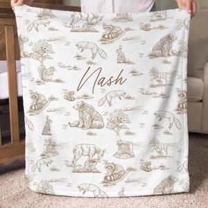 May include: White baby blanket with a brown woodland animal pattern, including bears, foxes, and raccoons. The name "Nash" is written in brown script across the blanket. The blanket is held up by a person.