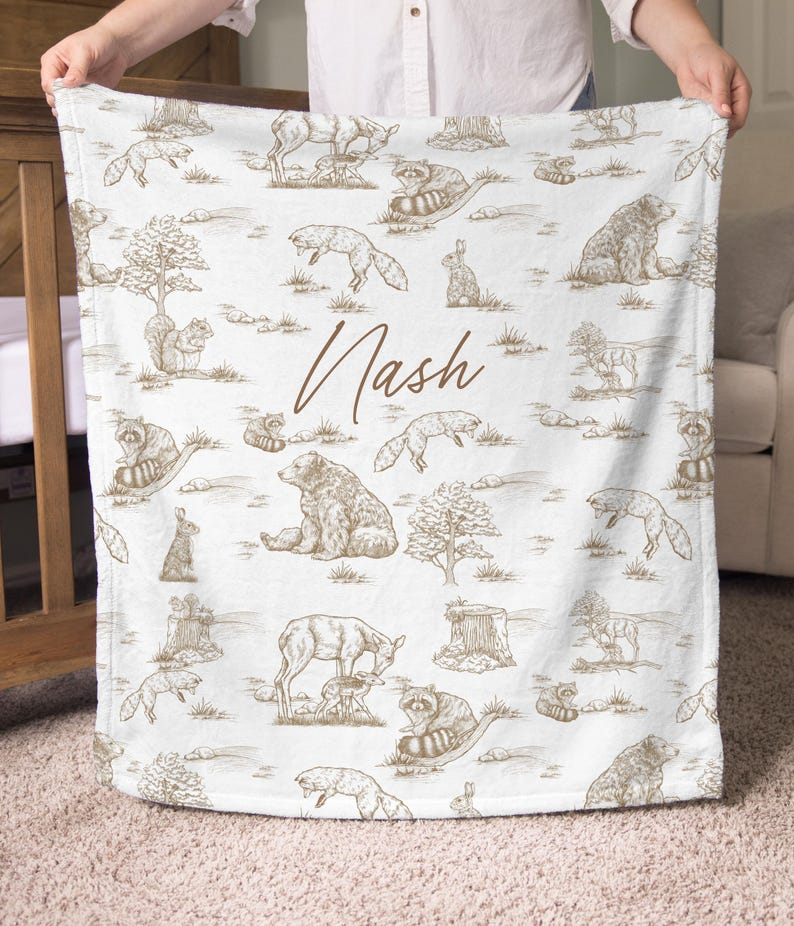 May include: White baby blanket with a brown woodland animal pattern, including bears, foxes, and raccoons. The name "Nash" is written in brown script across the blanket. The blanket is held up by a person.