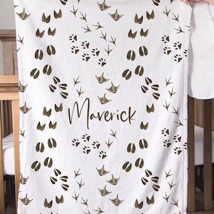 May include: White baby blanket with a repeating pattern of animal tracks in brown. The name "Maverick" is printed in a cursive font in the center of the blanket. The blanket is held up by a person.