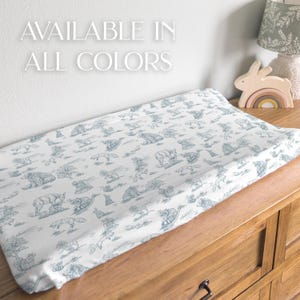 May include: A white changing pad cover with a blue animal print featuring bears, foxes, and other woodland creatures. The text "AVAILABLE IN ALL COLORS" is displayed in white. The pad is on a wooden dresser.