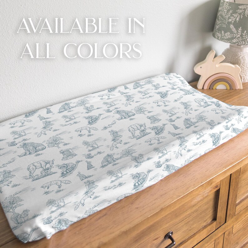 May include: A white changing pad cover with a blue animal print featuring bears, foxes, and other woodland creatures. The text "AVAILABLE IN ALL COLORS" is displayed in white. The pad is on a wooden dresser.
