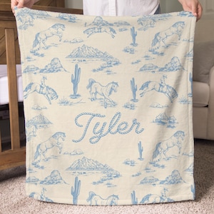 Puede incluir: Una manta color crema con un patr&oacute;n azul de tem&aacute;tica occidental. El dise&ntilde;o incluye caballos, cactus, monta&ntilde;as y un vaquero montando a caballo. El nombre "Tyler" est&aacute; escrito en cursiva.