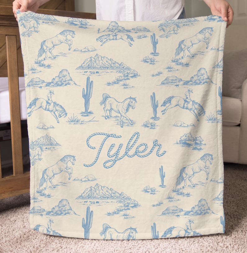 Puede incluir: Una manta color crema con un patr&oacute;n azul de tem&aacute;tica occidental. El dise&ntilde;o incluye caballos, cactus, monta&ntilde;as y un vaquero montando a caballo. El nombre "Tyler" est&aacute; escrito en cursiva.