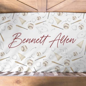 May include: A white crib sheet featuring a repeating pattern of baseballs, wooden bats, and baseball diamonds with arrows, centered with the name Bennett Allen written in a large, dark red cursive script across the middle of the fabric.