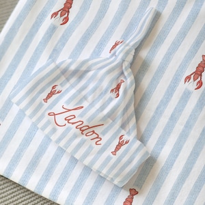Puede incluir: Un conjunto de gorro y manta para bebé con un estampado de rayas azules y blancas. El conjunto está decorado con ilustraciones de langostas rojas. El nombre "Lardon" está escrito en rojo en el gorro.