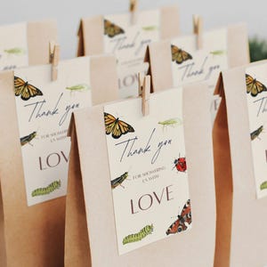 May include: Brown paper gift bags with tags that read "Thank you" and "Love." The tags feature watercolor illustrations of butterflies, insects, and caterpillars. The tags are attached with small wooden clothespins.