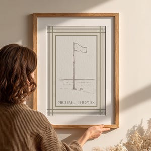 May include: A framed print featuring a golf course scene with a flag on a pole and a golf ball. The artwork is in a minimalist style with the name "MICHAEL THOMAS" printed at the bottom. The frame is made of wood.
