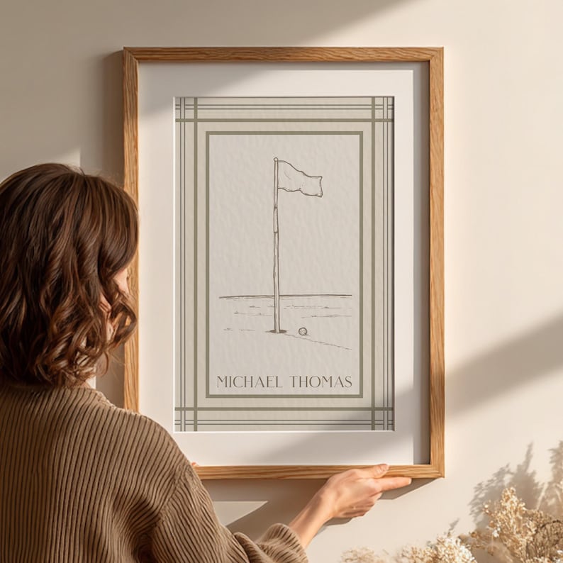 May include: A framed print featuring a golf course scene with a flag on a pole and a golf ball. The artwork is in a minimalist style with the name "MICHAEL THOMAS" printed at the bottom. The frame is made of wood.