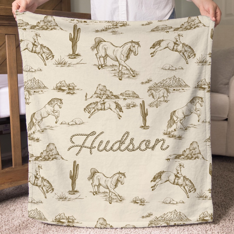 Puede incluir: Una manta color crema con un patr&oacute;n repetido de caballos, jinetes, cactus y monta&ntilde;as en marr&oacute;n. El nombre "Hudson" est&aacute; escrito en una fuente de guion. La manta est&aacute; sostenida por una persona.
