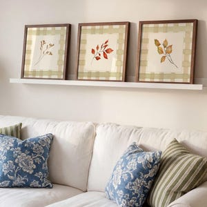 May include: Three square framed wall art pieces with dark wood frames, each featuring a botanical illustration of leaves on a cream background with a light green and white checkered border, displayed on a white floating shelf above a sofa.