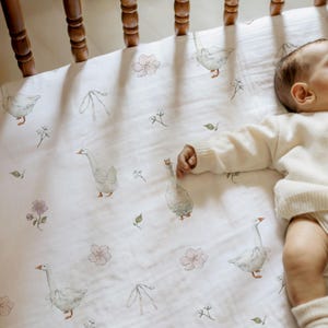 Puede incluir: Sábana blanca para cuna de bebé con un estampado de acuarela de gansos, flores rosas y hojas verdes. La sábana está hecha de una tela suave y transpirable. Un bebé está acostado en la sábana.
