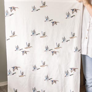Peut inclure: Un textile rectangulaire blanc orn&eacute; d'un motif r&eacute;p&eacute;titif de canards colverts en plein vol. Les canards sont repr&eacute;sent&eacute;s avec un plumage brun, vert et bleu, dispers&eacute;s sur le tissu dans diverses orientations, cr&eacute;ant un imprim&eacute; animalier coh&eacute;rent.