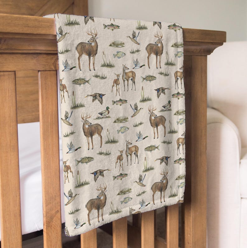 May include: A beige blanket with a repeating pattern of deer, ducks, fish, and reeds. The deer are brown with antlers, the ducks are in flight, and the fish are green. The blanket is draped over a wooden crib.