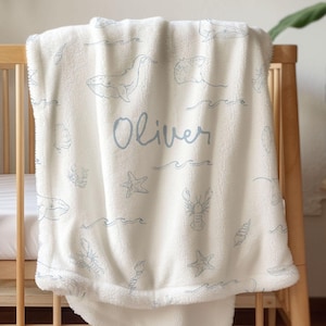 Könnte beinhalten: Eine weiche, weiße Fleecedecke mit einem wiederkehrenden Muster aus hellblauen Strichzeichnungen von Walen, Seesternen, Hummern, Muscheln und Meereswellen. Der Name Oliver ist mittig in einer passenden hellblauen Schreibschrift aufgedruckt.