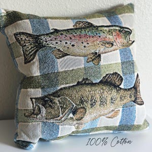 May include: A square throw pillow featuring a woven tapestry design of two fish against a blue, green, and white checkered background. The top fish is a rainbow trout with speckled markings, and the bottom fish is a bass. The text 100% Cotton is visible.