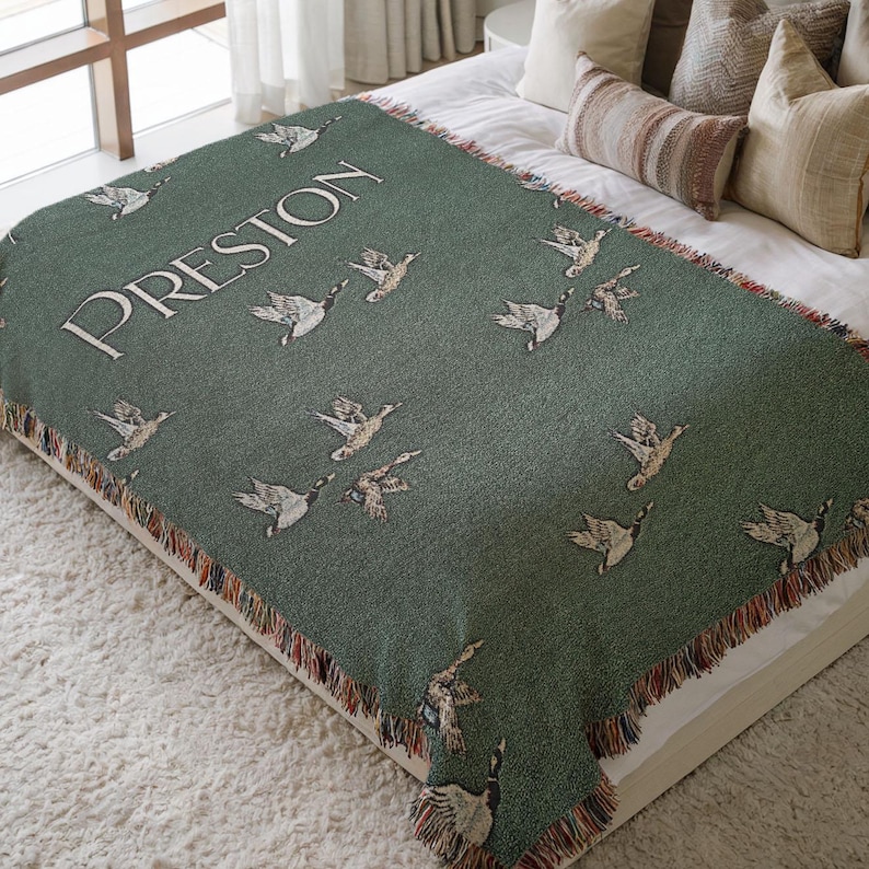 May include: A green woven throw blanket with a fringe border features a pattern of flying ducks and the name "Preston" woven in white.