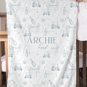 K&ouml;nnte beinhalten: Wei&szlig;es Babydecke mit einem Golf-Motiv in Hellblau. Das Design umfasst Golfwagen, Golftaschen, Golfb&auml;lle und Golfschl&auml;ger. Der Name "ARCHIE Ford" ist in der Mitte der Decke aufgedruckt.