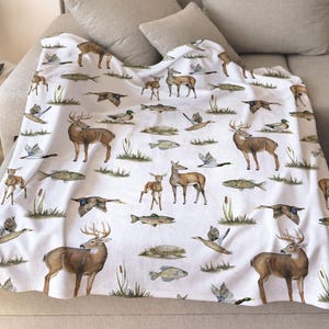 May include: White blanket featuring a repeating pattern of deer, ducks, and fish in shades of brown, green, and gray. The wildlife illustrations are scattered across the blanket, creating a nature-themed design. The blanket is draped over a beige sofa.