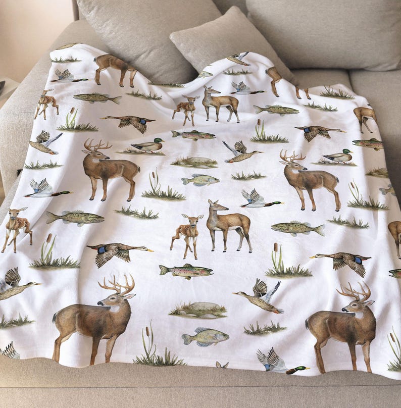 May include: White blanket featuring a repeating pattern of deer, ducks, and fish in shades of brown, green, and gray. The wildlife illustrations are scattered across the blanket, creating a nature-themed design. The blanket is draped over a beige sofa.