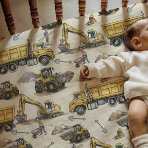 May include: A beige cot sheet with a repeating pattern of yellow construction vehicles, including dump trucks, excavators, and front-end loaders. The design includes illustrations of construction workers, shovels, and dirt mounds.