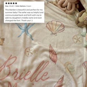 May include: Cream-colored baby blanket with watercolor seashell and starfish designs. The blanket measures 30x40 inches and features the name "Brielle" in pink script. The blanket is decorated with various sea-themed illustrations.