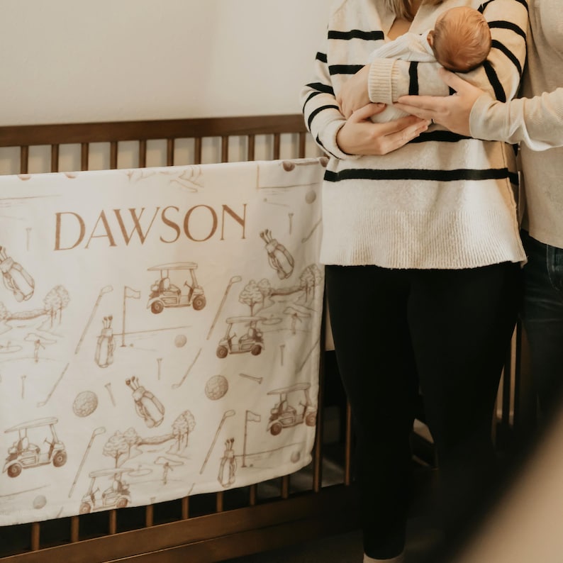 以下が含まれることがあります： 白いベビーブランケット。ゴルフコースのデザインと茶色の文字で「DAWSON」と書かれています。ブランケットはベビーベッドに掛けられています。画像には、赤ちゃんを抱いている人も写っています。