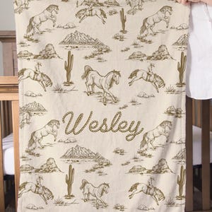 Puede incluir: Una manta beige con un patr&oacute;n repetido de caballos, vaqueros, cactus y monta&ntilde;as en marr&oacute;n. El nombre "Wesley" est&aacute; escrito en cursiva en el centro. La manta est&aacute; sostenida por una persona.