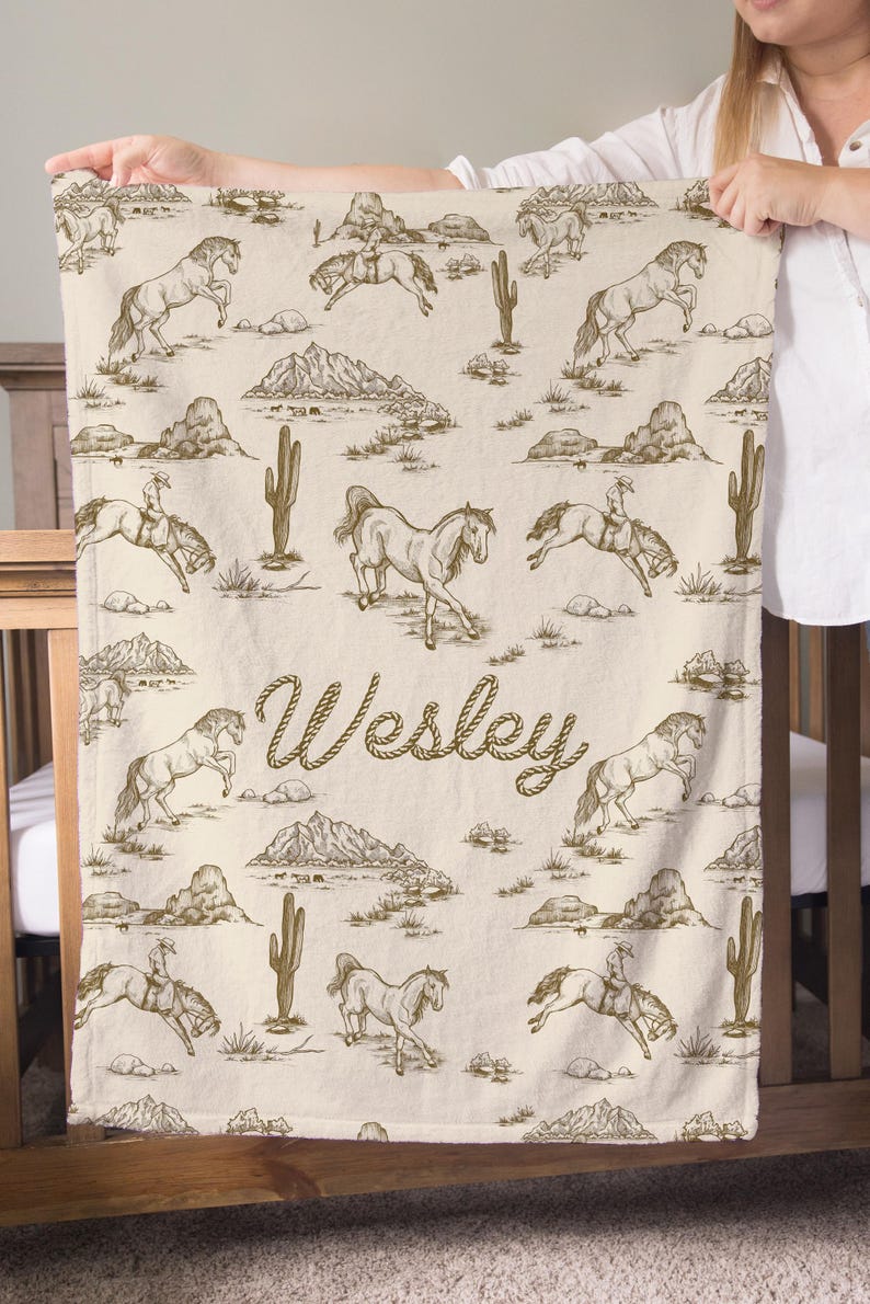 Puede incluir: Una manta beige con un patr&oacute;n repetido de caballos, vaqueros, cactus y monta&ntilde;as en marr&oacute;n. El nombre "Wesley" est&aacute; escrito en cursiva en el centro. La manta est&aacute; sostenida por una persona.