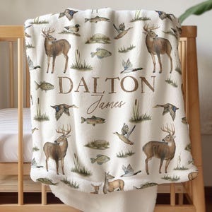 May include: A cream-colored baby blanket with a repeating pattern of deer, ducks, fish, and reeds. The blanket features the name "Dalton James" in brown lettering. The blanket is draped over a wooden crib.