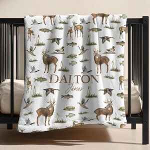May include: White blanket with a repeating pattern of deer, fish, ducks, and cattails. The blanket has the name "Dalton James" printed in brown script. The blanket is draped over a black crib.