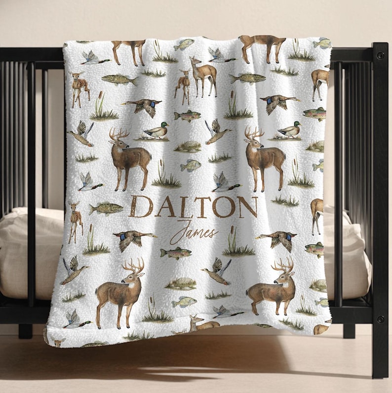 May include: White blanket with a repeating pattern of deer, fish, ducks, and cattails. The blanket has the name "Dalton James" printed in brown script. The blanket is draped over a black crib.