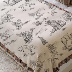 May include: Cream-colored throw blanket with a black and white forest animal design. The blanket features a repeating pattern of bears, foxes, and other woodland creatures. The edges are finished with a fringe.