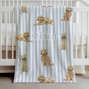 May include: A baby blanket with a blue and white striped pattern. The blanket features illustrations of golden doodle dogs in various poses. The name "Nolan" is printed in light blue on the blanket.