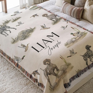 May include: A cream-coloured woven throw blanket featuring a repeating pattern of hunting dogs and ducks in a marsh setting. The centre displays the name LIAM Joseph in black lettering. The edges are finished with a multi-coloured fringe trim.