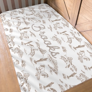 Puede incluir: Una sábana bajera blanca para cuna con un patrón repetido de vaqueros, caballos, cactus y otras ilustraciones de temática occidental en tonos marrones. El nombre "Brooks" está escrito con letras de cuerda.